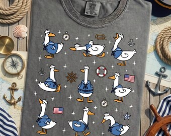 Könnte beinhalten: Dunkelgraues T-Shirt mit einem Muster von Cartoon-Seemannsgänsen in blau-weißen Matrosenoutfits. Das Design umfasst nautische Elemente wie Anker, Kompasse und die amerikanische Flagge. Das Shirt hat eine entspannte Passform.