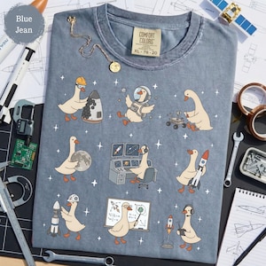 May include: A blue jean colored t-shirt featuring a graphic print of white geese in space. The geese are depicted wearing astronaut helmets, working at computer consoles, holding rockets, and examining the moon, all surrounded by small white stars.