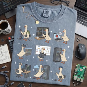 May include: A light blue t-shirt featuring a cartoon goose in various tech-related scenarios, such as coding and server management. The shirt includes the text "COMFORT COLORS" and size information. A coffee mug with "CODE FUEL" is also visible.