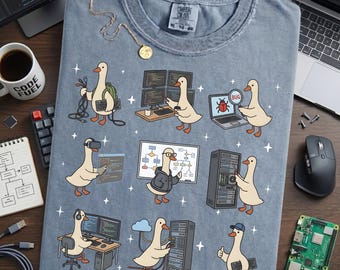 May include: A light blue t-shirt featuring a cartoon goose in various tech-related scenarios, such as coding and server management. The shirt includes the text "COMFORT COLORS" and size information. A coffee mug with "CODE FUEL" is also visible.
