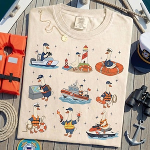 May include: Cream-colored t-shirt featuring cartoon geese in Coast Guard uniforms. The design includes boats, a lighthouse, and life preservers. A gold necklace and a life vest are also visible.