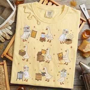 May include: Yellow Comfort Colors t-shirt featuring cartoon geese dressed as beekeepers. The geese are shown working with honeycombs, hives, and other beekeeping equipment. A gold necklace is draped on the shirt.