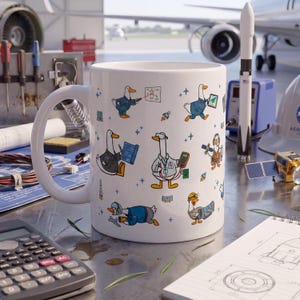 May include: A white ceramic mug featuring a repeating pattern of cartoon ducks dressed as aerospace engineers and technicians, performing tasks like using laser pointers, holding blueprints, operating multimeters, and wearing safety gear.