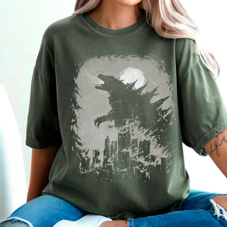 May include: Olive green t-shirt featuring a Godzilla graphic. The design shows Godzilla silhouetted against a full moon and city skyline. The graphic is in shades of gray and white, creating a vintage, distressed look.