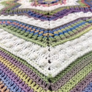 Puede incluir: Primer plano de una manta de ganchillo hecha a mano. La manta presenta un dise&ntilde;o geom&eacute;trico con una variedad de colores, incluyendo blanco, morado, verde, azul y amarillo. La textura es visible, con puntadas en relieve y un patr&oacute;n detallado.