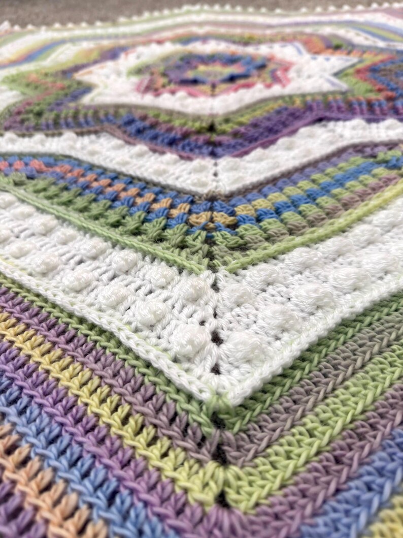 Puede incluir: Primer plano de una manta de ganchillo hecha a mano. La manta presenta un dise&ntilde;o geom&eacute;trico con una variedad de colores, incluyendo blanco, morado, verde, azul y amarillo. La textura es visible, con puntadas en relieve y un patr&oacute;n detallado.