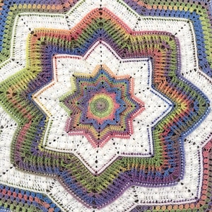 Puede incluir: Una manta de ganchillo hecha a mano y colorida con un dise&ntilde;o de estrella. La manta presenta una estrella central en tonos de rosa, verde y azul, rodeada de formas de estrella m&aacute;s grandes en blanco y colores del arco&iacute;ris. El intrincado patr&oacute;n es visible.