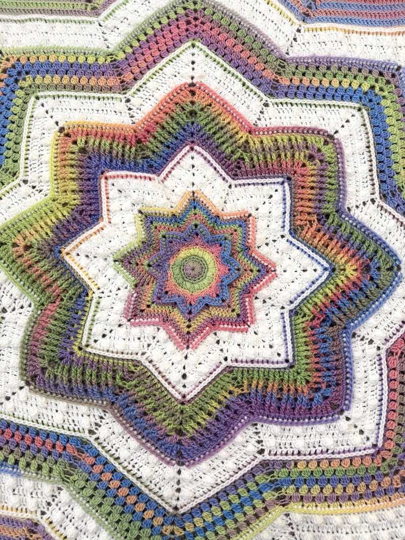 Puede incluir: Una manta de ganchillo hecha a mano y colorida con un dise&ntilde;o de estrella. La manta presenta una estrella central en tonos de rosa, verde y azul, rodeada de formas de estrella m&aacute;s grandes en blanco y colores del arco&iacute;ris. El intrincado patr&oacute;n es visible.