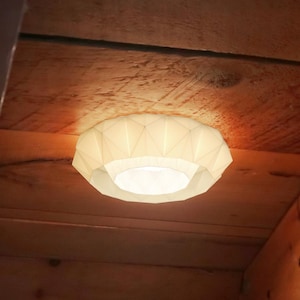 May include: A white, geometric ceiling light is fixed to a wooden ceiling. The light emits a warm glow, illuminating the surrounding wooden beams and planks. The fixture's design is modern and minimalist, suitable for various interior styles.