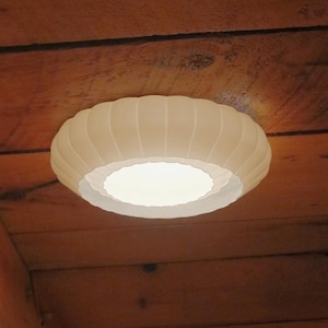 May include: A white, round ceiling light fixture with a ribbed design, mounted on a wooden ceiling. The light emits a soft glow, illuminating the surrounding wooden beams and planks. The fixture's design adds a touch of elegance to the rustic setting.
