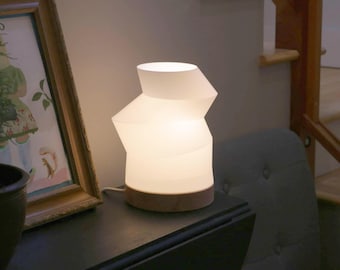 May include: A contemporary white table lamp with a distinctive geometric shape. The lamp features a wooden base and provides a gentle, warm light. It sits on a dark surface, beside a framed picture and a potted plant.