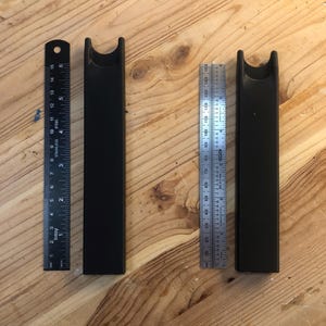 Könnte beinhalten: Zwei schwarze, rechteckige Objekte mit gewölbter Oberseite, neben zwei Linealen. Ein Lineal ist in Zoll, das andere in Zentimetern markiert. Die Objekte scheinen aus einem festen Material zu bestehen. Der Hintergrund ist eine hellfarbige Holzoberfläche.