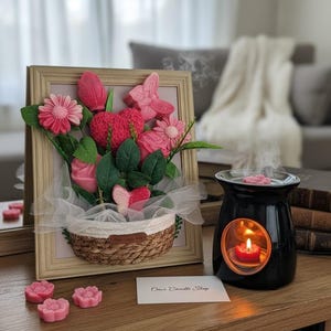 Peut inclure: Composition florale encadrée avec des fleurs roses, des papillons et des feuilles vertes dans un panier en osier. Un diffuseur d'huiles essentielles noir avec une bougie allumée est à côté. Le texte "Omis Candle Shop" est sur une carte.