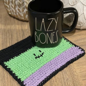 May include: A black ceramic mug with the words "LAZY BONES" in white lettering. The mug sits on a handmade crochet coaster with black, green, and purple stripes and a stitched face. Decorative gourds are in the background.
