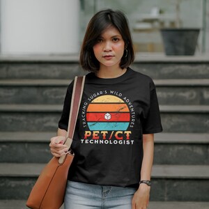 Puede incluir: Una camiseta negra con un diseño gráfico de estilo vintage. El gráfico presenta una puesta de sol con el texto "Tracking Sugar's Wild Adventures" y "PET/CT Technologist".