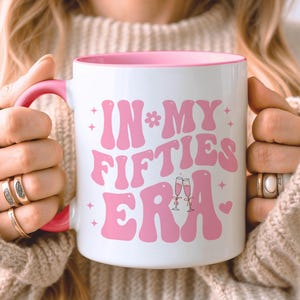 May include: White ceramic mug with a pink interior and handle. The mug features the phrase "IN MY FIFTIES ERA" in pink retro font, with star and flower accents. Two champagne glasses and a heart are also included in the design.