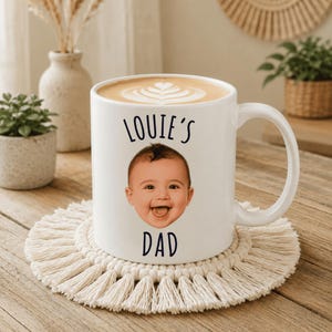 May include: A white ceramic mug filled with a latte featuring a heart-shaped foam design. The mug displays a photographic print of a baby's face centered between the black text "LOUIE'S" above and "DAD" below, with a curved handle on the right.