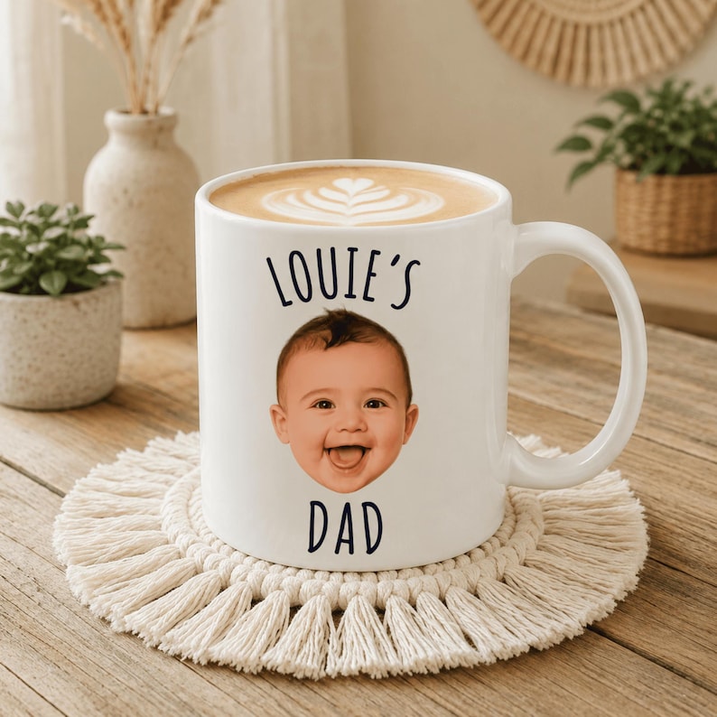 May include: A white ceramic mug filled with a latte featuring a heart-shaped foam design. The mug displays a photographic print of a baby's face centered between the black text "LOUIE'S" above and "DAD" below, with a curved handle on the right.