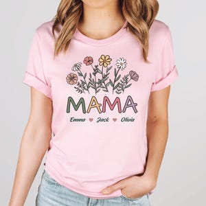 May include: Pink t-shirt featuring a floral design and the word "MAMA" in colourful letters. Below the word "MAMA" are the names "Emma", "Jack", and "Olivia" with hearts between each name.
