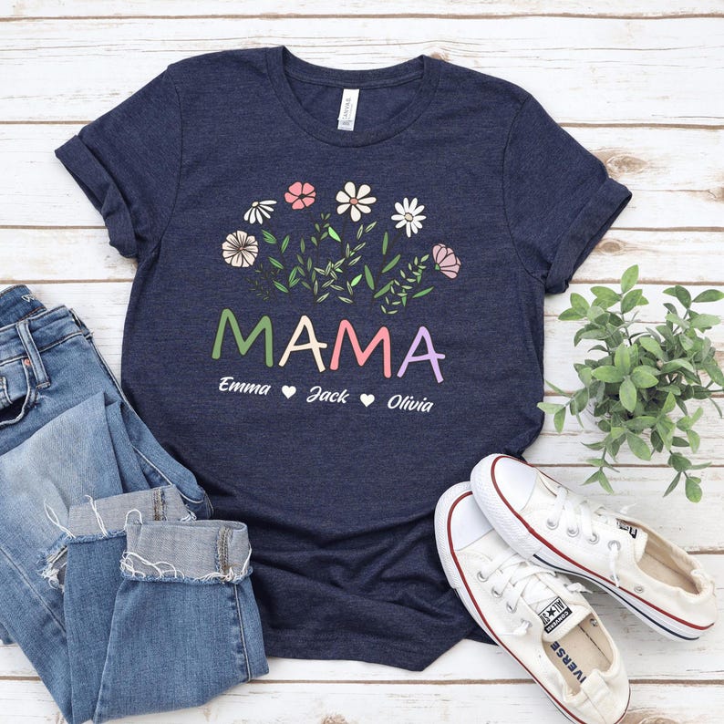 May include: A navy blue t-shirt with a floral design and the word "MAMA" in colourful letters. Below the word "MAMA" are the names "Emma", "Jack", and "Olivia" with hearts between each name.