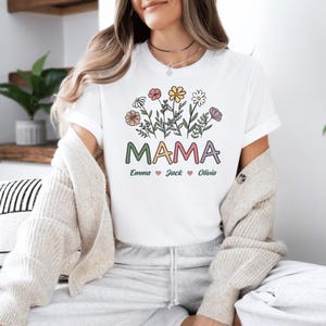 May include: White t-shirt featuring a floral design and the word "MAMA" in colourful letters. Below the word "MAMA" are the names "Emma", "Jack", and "Olivia" with hearts between each name.