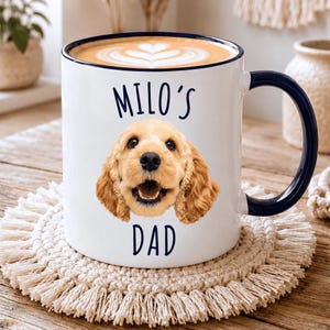 Può includere: Una tazza in ceramica bianca con bordo e manico blu scuro, caratterizzata da una stampa fotografica del muso di un cane spaniel color oro al centro. Il testo "MILO'S" è stampato in blu scuro sopra il cane e "DAD" al di sotto.