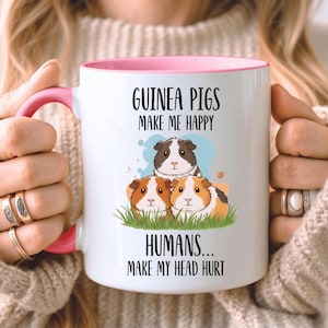 May include: White ceramic mug with a pink interior and handle, featuring a graphic of three guinea pigs and the text "GUINEA PIGS MAKE ME HAPPY HUMANS... MAKE MY HEAD HURT".