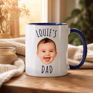 May include: A white ceramic mug with a dark blue interior and handle. The front features the text "LOUIE'S" above a photographic print of a smiling baby's face, with the word "DAD" printed in dark blue lettering directly below the face.