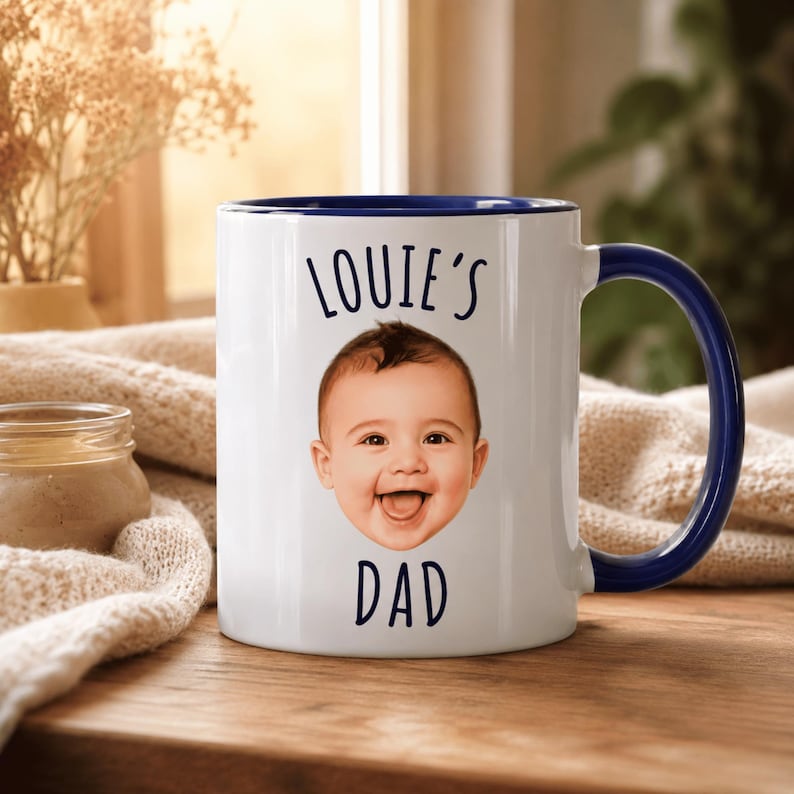 May include: A white ceramic mug with a dark blue interior and handle. The front features the text "LOUIE'S" above a photographic print of a smiling baby's face, with the word "DAD" printed in dark blue lettering directly below the face.