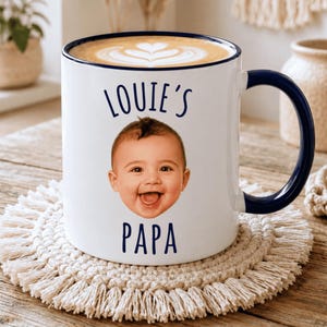 May include: A white ceramic mug with a dark blue rim and handle, featuring a photographic print of a baby's face in the center. The text "LOUIE'S" is printed in dark blue above the baby's face, and the word "PAPA" is printed below it.