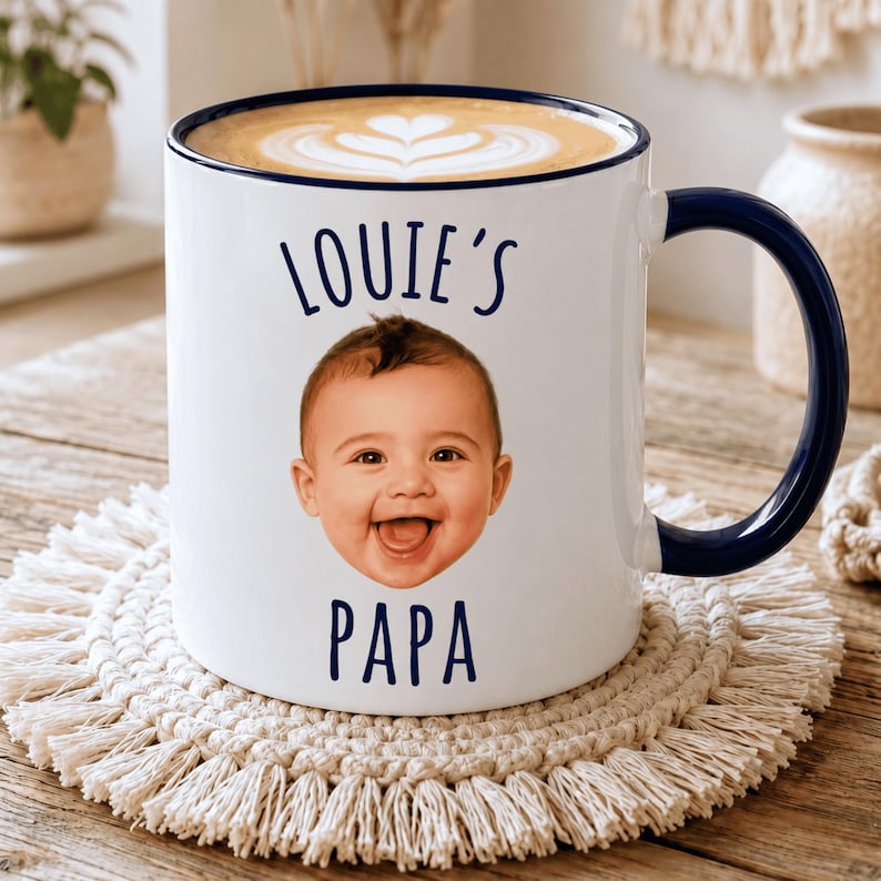 May include: A white ceramic mug with a dark blue rim and handle, featuring a photographic print of a baby's face in the center. The text "LOUIE'S" is printed in dark blue above the baby's face, and the word "PAPA" is printed below it.