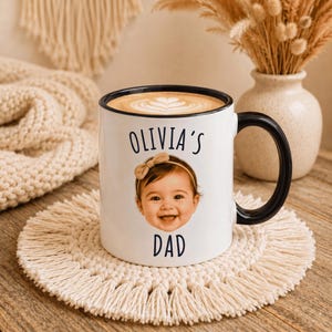 May include: A white ceramic mug with a black rim and handle, featuring a printed photograph of a baby wearing a tan bow headband. The text "OLIVIA'S" is printed in black above the baby's face, and the word "DAD" is printed in black below it.