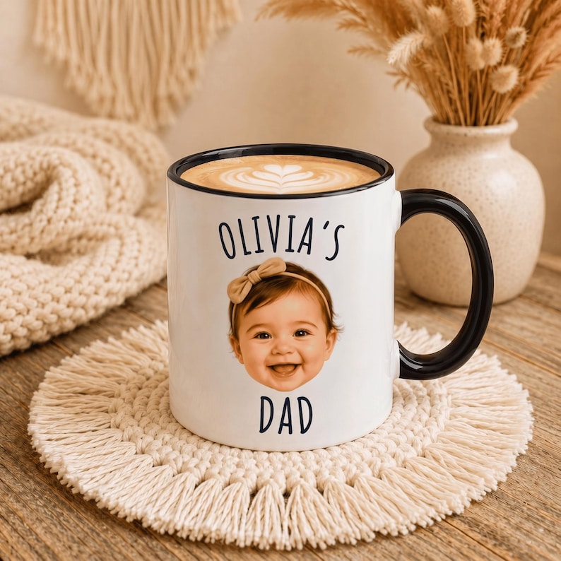 May include: A white ceramic mug with a black rim and handle, featuring a printed photograph of a baby wearing a tan bow headband. The text "OLIVIA'S" is printed in black above the baby's face, and the word "DAD" is printed in black below it.