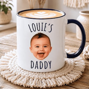 May include: A white ceramic mug with a dark blue rim and handle, featuring a photographic print of a smiling baby's face centred between the dark blue text that reads LOUIE'S on the top and DADDY on the bottom, filled with a latte.