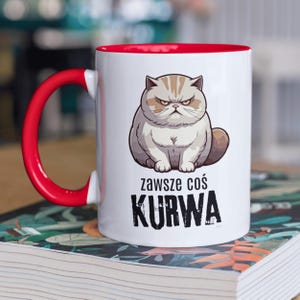 May include: White ceramic mug with a red rim and handle. The mug features a cartoon illustration of an angry cat with the text "zawsze coś KURWA" printed below.