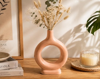 May include: A matte peach-colored ceramic vase with a circular, donut-shaped body and a narrow neck. The vase is filled with a dried floral arrangement consisting of wheat stalks, small white flowers, and green eucalyptus leaves.