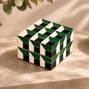 May include: A square box featuring a geometric pattern of alternating black, white, and dark green shapes. The lid and base are aligned, creating a continuous three-dimensional cube effect across the exterior surfaces of the container.
