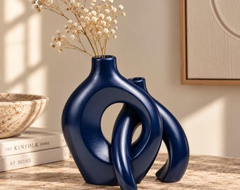 May include: A dark blue ceramic vase with an abstract, interconnected double-loop design and a smooth finish. The vase features two narrow openings at the top, one of which holds a delicate sprig of dried white baby's breath flowers.