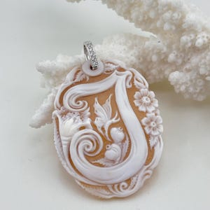 Pode incluir: Pingente oval com um design floral e de rolagem esculpido em branco sobre um fundo bege. O pingente tem uma argola prateada com pequenas pedras transparentes. O pingente é provavelmente feito de concha ou material semelhante.