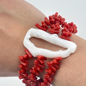 Könnte beinhalten: Ein rotes Korallenarmband mit einem weißen, länglichen Mittelstück. Das Armband besteht aus mehreren Strängen kleiner, roter Korallenstücke. Das weiße Mittelstück hat eine unregelmäßige, organische Form. Das Armband wird am Handgelenk getragen.