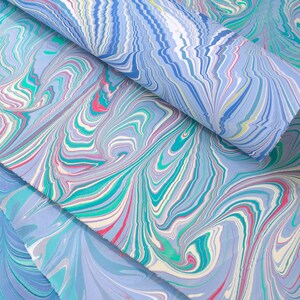 May include: Close-up of marbled paper sheets in shades of blue, teal, pink, and white. The paper has a swirling, organic pattern, with a rolled sheet on top. The paper is likely used for art, crafts, or stationery.