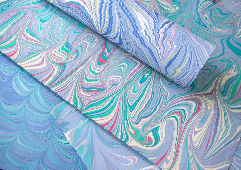 May include: Close-up of marbled paper sheets in shades of blue, teal, pink, and white. The paper has a swirling, organic pattern, with a rolled sheet on top. The paper is likely used for art, crafts, or stationery.