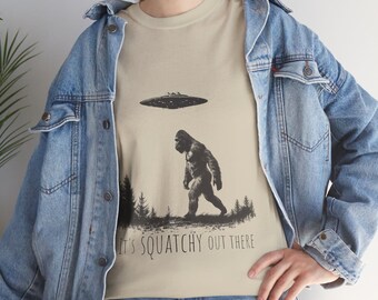 Könnte beinhalten: Ein beiges T-Shirt mit einer schwarz-weißen Grafik von Bigfoot, der auf ein UFO zugeht. Der Text "It's Squatchy Out There" ist unter der Grafik aufgedruckt.