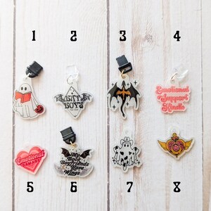 May include: Eight different clear acrylic charms with various designs. The charms are attached to black and gold clips. The designs include a ghost reading a book, a bat with the text "Bat Boys Fan Club", a black dragon, a ghost with a cowboy hat, a heart with the text "Emotional Damage", a bat with the text "My Book Boyfriends Have Wings", a heart with wings and a crown, and a pink heart with the text "Emotional Support Kindle".