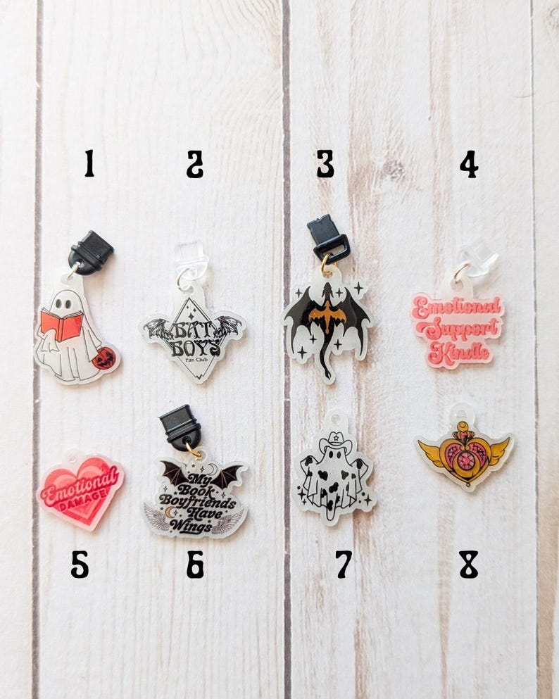 May include: Eight different clear acrylic charms with various designs. The charms are attached to black and gold clips. The designs include a ghost reading a book, a bat with the text "Bat Boys Fan Club", a black dragon, a ghost with a cowboy hat, a heart with the text "Emotional Damage", a bat with the text "My Book Boyfriends Have Wings", a heart with wings and a crown, and a pink heart with the text "Emotional Support Kindle".