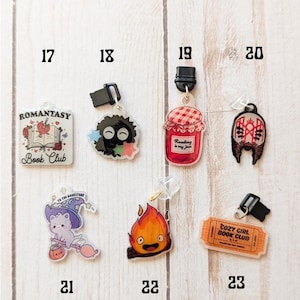 May include: A set of 10 colorful book charms featuring various designs, including a book club logo, a jar of jam with the text "Reading is my jam", a cat with a witch hat, and a fire spirit. The charms are made of clear acrylic and have a black clip on the top.