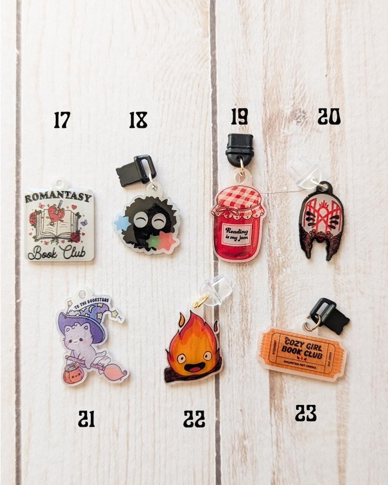 May include: A set of 10 colorful book charms featuring various designs, including a book club logo, a jar of jam with the text "Reading is my jam", a cat with a witch hat, and a fire spirit. The charms are made of clear acrylic and have a black clip on the top.