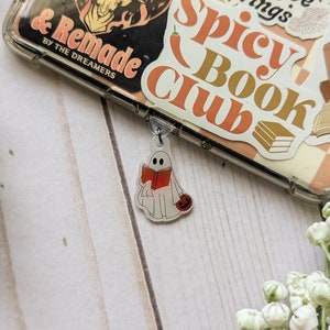 May include: A white ghost holding a red book and a small orange pumpkin. The ghost is a charm attached to a clear phone case with stickers that say "Spicy Book Club" and "& Remade by the Dreamers".