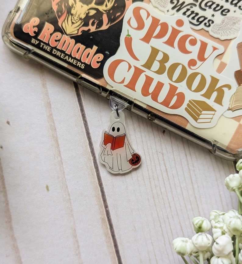 May include: A white ghost holding a red book and a small orange pumpkin. The ghost is a charm attached to a clear phone case with stickers that say "Spicy Book Club" and "& Remade by the Dreamers".