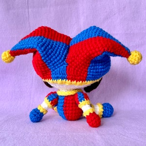 May include: A handmade crochet jester doll in red, blue, yellow, and white. The doll features a traditional jester hat with bells, a colorful outfit, and black eyes. The doll is sitting on a purple surface.
